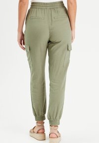 Pantalon cargo vert olive avec une ceinture élastique, deux poches arrière, et des poignets fuselés. Dispose de poches latérales utilitaires. Texture de tissu douce.