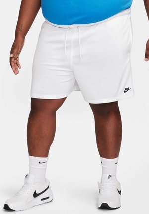 Homme portant un short blanc Nike, des chaussettes blanches Nike et des baskets blanches Nike Air, debout devant un fond uni.