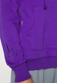Manche de sweat à capuche violette avec des lettres brodées "A|L", une poche latérale zippée et une poche kangourou sur le torse avant au-dessus d’un pantalon gris.