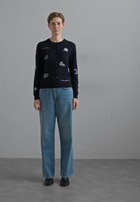 Cardigan blu navy con chiusura a bottoni, caratterizzato da design ricamati, abbinato a jeans a gamba larga blu chiaro e stivali alla caviglia neri.