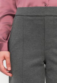 Pantaloni grigi tessuti con una consistenza liscia e una vestibilità sartoriale, con vita alta e tasche laterali. Abbinati a una blusa in satin rosa.
