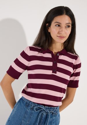 Vrouw met lang donker haar die een roze en bordeaux gestreept poloshirt met korte mouwen draagt en een hoge taille blauwe spijkerbroek met een gestrikte riem.