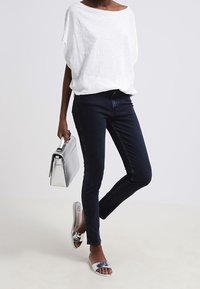 Una mujer se encuentra de pie con confianza, vistiendo una blusa blanca suelta, jeans oscuros y sandalias planas plateadas, sosteniendo un bolso metálico.