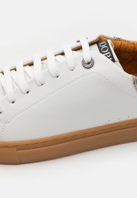 Zapatilla de cuero blanca con suela de goma texturada en tono marrón, seis ojales blancos y forro interior marrón. Presenta un detalle de logo circular en el lateral.