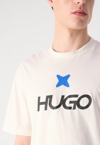 Cream-colored cotton t-shirt featuring a blue star graphic above the black "HUGO" logo. Short sleeves and a crew neckline. Smooth texture.
