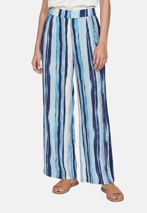 Femme portant un pantalon large à rayures verticales bleues et blanches, assorti à des sandales ouvertes couleur beige et un haut blanc.