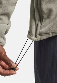 Fleecejacke in Hellgrau mit einer Hand, die einen Kordelzug am Saum anpasst. Verfügt über eine weiche Textur und Bündchendetails mit einem schwarzen Kordel.