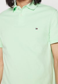 Světle zelená polokošile s krátkým rukávem a límečkem, na levé straně hrudi je malé obdélníkové logo v červené, bílé a modré barvě.
