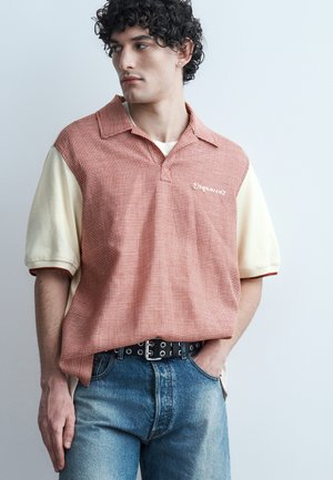 Jeune homme aux cheveux bouclés portant une chemise bicolore rouge et crème à manches courtes et un jean bleu, regardant vers sa gauche avec une main dans la poche.