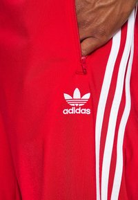 Röd träningsjacka med en vit dragkedja; har Adidas-logga i vitt och tre vertikala vita ränder på axeln. Slät textur.