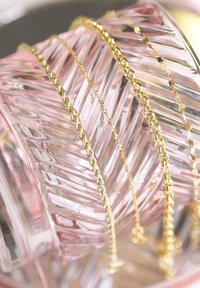 Three gold chains with varying designs resting on a textured pink glass surface. Chains include twisted, linked, and flat styles.