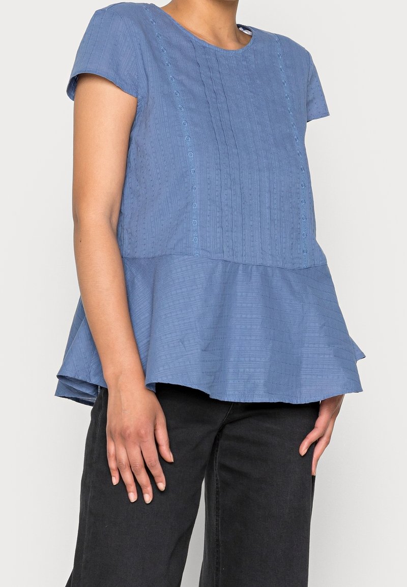 Haut peplum bleu avec des rayures texturées, manches courtes, col rond, et des détails de boutons sur le devant. Associé à un pantalon noir.