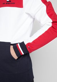 Czerwono-biało-granatowy sweatshirt z okrągłym dekoltem, z wielokolorowym wzorem w blokach, z ribbowymi ściągaczami i kieszenią na dolnej części przodu.