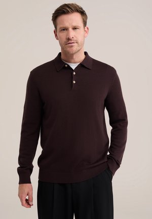 Homme portant un polo marron foncé à manches longues avec trois boutons, debout devant un fond clair uni.