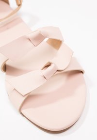 Sandale rose clair avec deux lanières en cuir nouées sur le dessus du pied et une semelle lisse sur fond blanc.