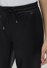 Sorte faux læder joggers med elastisk talje, der har en snor og metalender. Nærbillede af taljen og hånden.