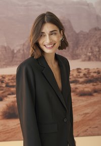 Juodas švarkas su pritaikytu sijonu, pasižymintis notch tipo apykakle ir vienu mygtuko uždarymu, dėvimas virš gilių iškirpės. Auksiniai geometriniai auskarai.