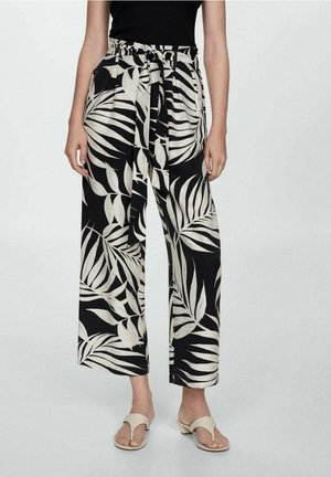 Woman wearing wide-leg black pants with large white tropical leaf print, paired with beige open-toe low-heeled sandals and a black top.