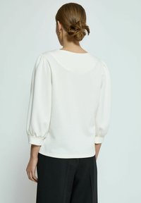 Witte lange mouwen top met opbollende schouders, ronde halslijn en een gladde textuur, gecombineerd met zwarte broek, gezien vanuit de achterkant.
