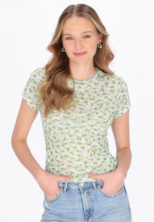 Junge Frau mit welligen blonden Haaren, die ein hellgrünes, gemustertes T-Shirt und blaue Jeans trägt, Hände in den Taschen, lächelnd vor weißem Hintergrund.