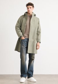Abrigo impermeable verde claro con capucha, de corte relajado, con botones a presión, usado sobre un suéter beige, combinado con jeans oscuros y zapatillas blancas.
