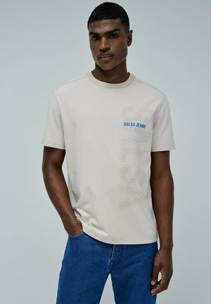 T-shirt beige à manches courtes avec un motif texturé et un logo "Salsa Jeans" bleu. Porté avec un jean en denim bleu, de coupe classique.