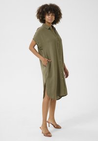Rochie cambrată, de culoare verde măslin, cu nasturi, mâneci scurte, buzunare laterale și tiv asimetric. Material texturat, cu o croială lejeră și detalii minime.