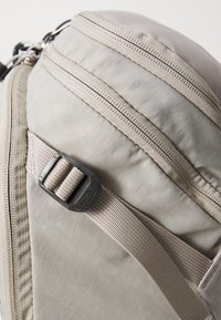 Light gray backpack featuring dual zippers, webbed strap with adjustable buckle, and textured fabric. Accents in darker gray.