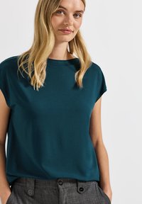 Top teal à manches courtes avec une coupe décontract�ée et un col rond. Texture lisse qui complète le pantalon gris à motifs.