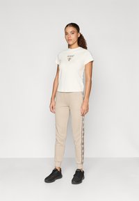 Camiseta blanca con un pequeño logotipo marrón, combinada con pantalones deportivos beige claros que presentan un patrón de logotipos a lo largo de los laterales. Zapatillas deportivas negras.