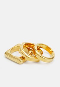 Tre anelli d'oro: uno rotondo, uno quadrato con bordi arrotondati e uno quadrato piatto. Finitura in metallo lucido, texture liscia e superfici lucidate.