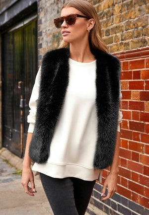 Gilet en fausse fourrure noire sur un sweat-shirt blanc, associé à un jean foncé. Le mannequin porte des lunettes de soleil en écaille dans un cadre urbain.