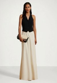 Femeie îmbrăcată cu o vestă neagră fără mâneci, cu nasturi, și o fustă lungă bej, ținând o poșetă cu tinte bej și neagră.