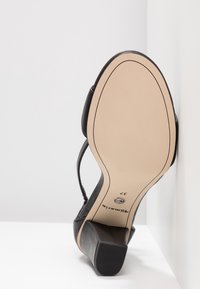 Sandale noire à talon avec semelle beige et taille 37 imprimée, présentée semelle vers le haut contre des murs et une surface blancs.