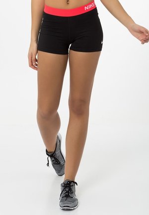 Vrouw draagt zwarte Nike short met rode tailleband en grijze sportschoenen, loopt vooruit tegen een witte achtergrond.