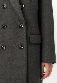 Manteau en laine double boutonnage gris foncé avec six boutons noirs et une poche à rabat, porté par une personne ayant une main visible sur le côté.