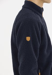 Laikalla fleece takki, jossa on korkea kaulus ja teksturoitu pinta. Varustettu nahkalaatalla ja vetoketjullisella sivutaskulla.