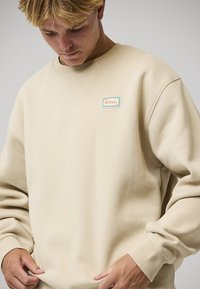 Sweat-shirt beige avec un col rond, des poignets côtelés, et une petite étiquette rectangulaire verte et blanche portant l'inscription « Rivvia ». Tissu doux et texturé.