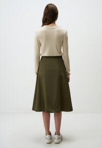Beige lange mouwen top, ronde halslijn, gecombineerd met een middellange olijfgroene rok. Witte sneakers zichtbaar aan de onderkant, gladde stof textuur.