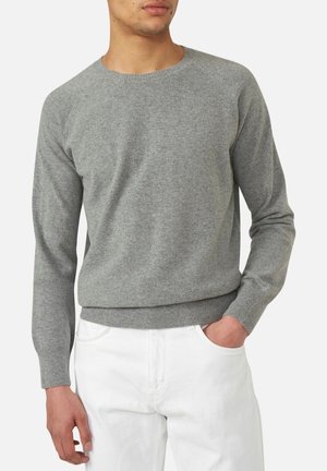 Uomo con maglione grigio aderente a maniche lunghe e pantaloni bianchi, con una mano in tasca, mostrato dal collo ai fianchi.