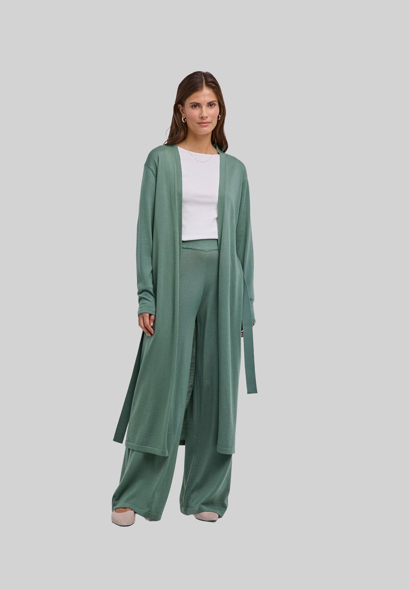 Donna che indossa pantaloni a gamba larga color salvia e un cardigan lungo sopra una maglietta bianca, in piedi davanti a uno sfondo grigio chiaro uniforme.