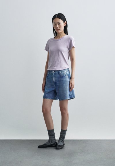 Mladá žena stojaca, oblečená v svetlofialovej krátkosleeve košeli, rifľových šortkách po kolená, sivých ponožkách a čiernych balerínach na jednoduchom pozadí.