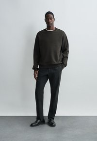 Sweatshirt verde scuro con collo a giro, polsini a costine, abbinato a pantaloni neri su misura e scarpe nere, posto su uno sfondo semplice.