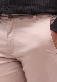 Κοντινό πλάνο ενός ατόμου που φοράει μπεζ παντελόνια με το χέρι στην τσέπη και μαύρο πουκάμισο από κορδούρα, μπαλωμένο  στη μέση.