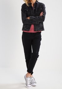 Giacca di jeans nera, t-shirt rossa, pantaloni neri e sneakers argentate con lacci. Il modello sta in piedi con le braccia conserte, mostrando un outfit a strati.