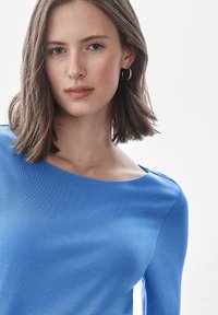 Blå, figurnära långärmad topp i mjukt tyg med vid halsringning och subtil textur. Modellen bär små silverringar.