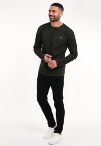 Maglietta verde oliva a maniche lunghe con scollatura rotonda e un piccolo logo sul petto, abbinata a pantaloni neri slim-fit e scarpe bianche.