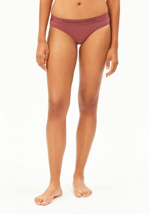 Femme debout pieds nus, portant un sous-vêtement taille basse mauve avec le logo ARMEDANGELS sur la ceinture, sur fond blanc.