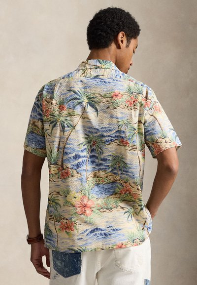 Muž nosi krátky rukáv havajskej košele s motívom tropických paliem, kvetov a vĺn oceánu, v kombinácii s bielymi roztrhanými krátkymi nohavicami.