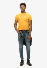 Orange bomulds t-shirt, blå jeans, ternet skjorte bundet om livet, sorte snørebåndsstøvler, sølv armbåndsur, afslappet design.